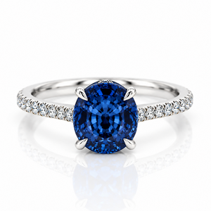 An oval-cut blue sapphire engagement ring with a silver-colored band accented by pavé-set diamonds, shown on a reflective white surface.
