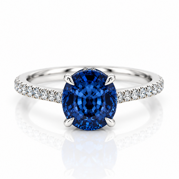 An oval-cut blue sapphire engagement ring with a silver-colored band accented by pavé-set diamonds, shown on a reflective white surface.