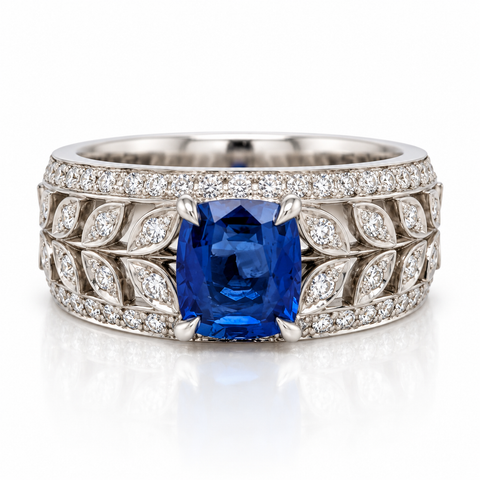 An Art Deco style diamond ring featuring a cushion-cut blue sapphire center stone, surrounded by a silver-toned leaf-pattern setting paved with small round diamonds, shown on a white background with a soft reflection.